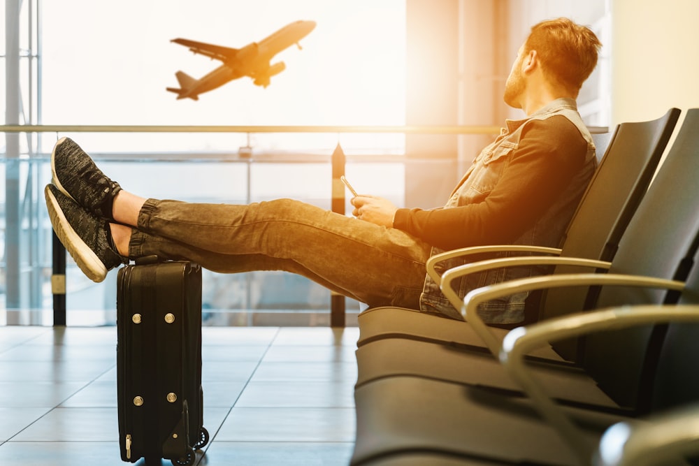 A traveller relaxing in an airport terminal with feet resting on a suitcase, watching a plane take off